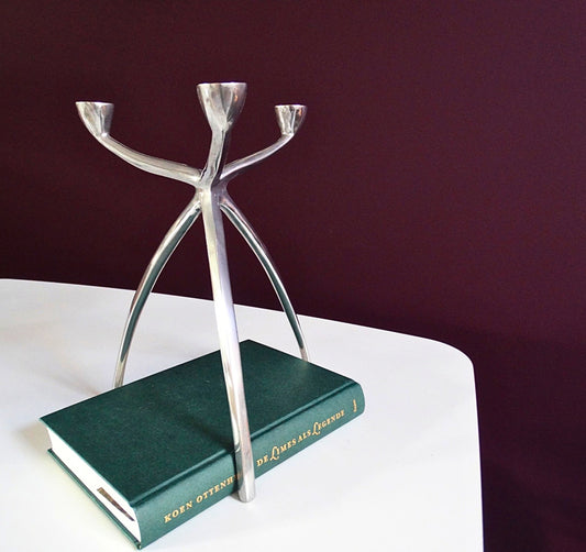 Space Age Aluminium Kandelaar voor drie kaarsen. Gefotografeerd op een witte tafel met een boek eronder als vergelijking voor grootte. Vintage van Atelier Aeris verkrijgbaar bij Draumr