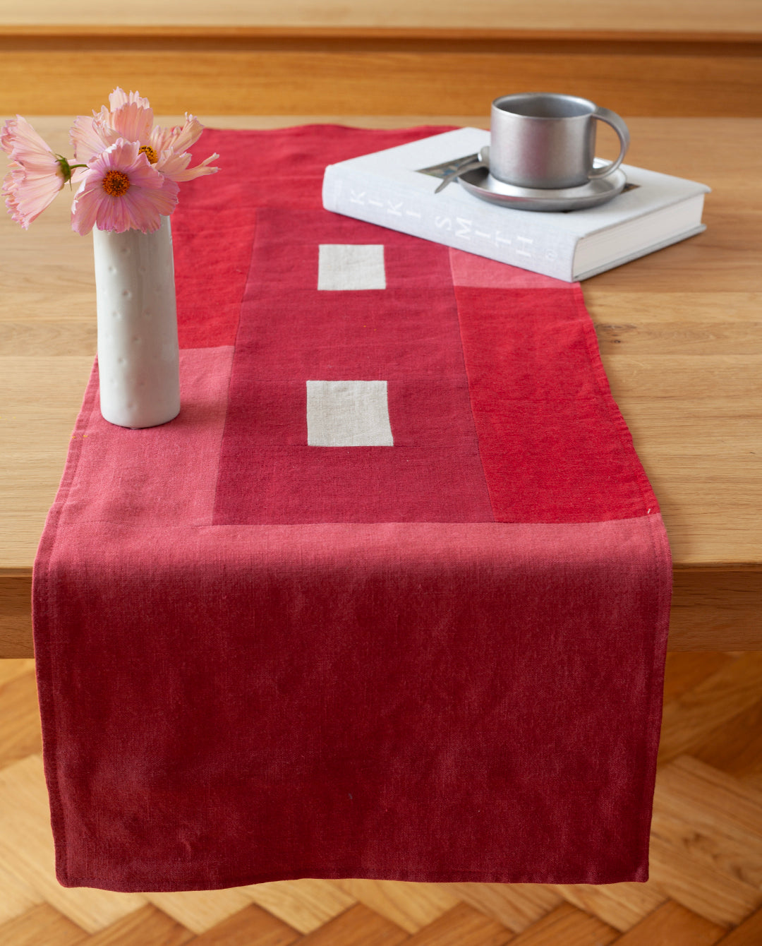 Een sfeervolle foto van een rode  tafelrunner ontworpen door Kathryn Clark, gedrapeerd over een houten tafel.