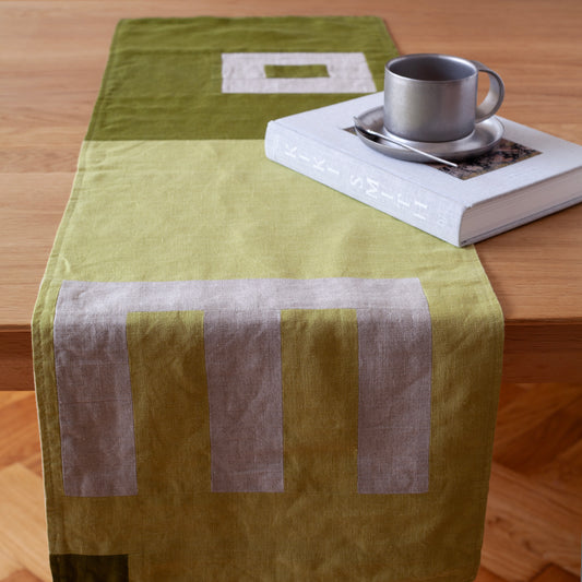 Een overzicht van een mos groene tafelrunner ontworpen door Kathryn Clark gelegd over een houten tafel met een boek en een koffiekopje
