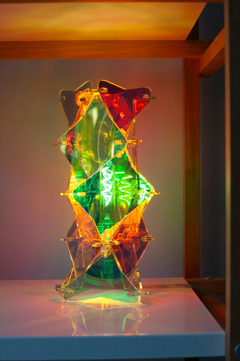 Een tafellamp in de kenmerkende irisiderende sfeer die je kent van Lux Vortex. De lamp is een tafelmodel en staat in een kast De lamp is nu verlicht en je ziet de draad in de lamp zitten.
